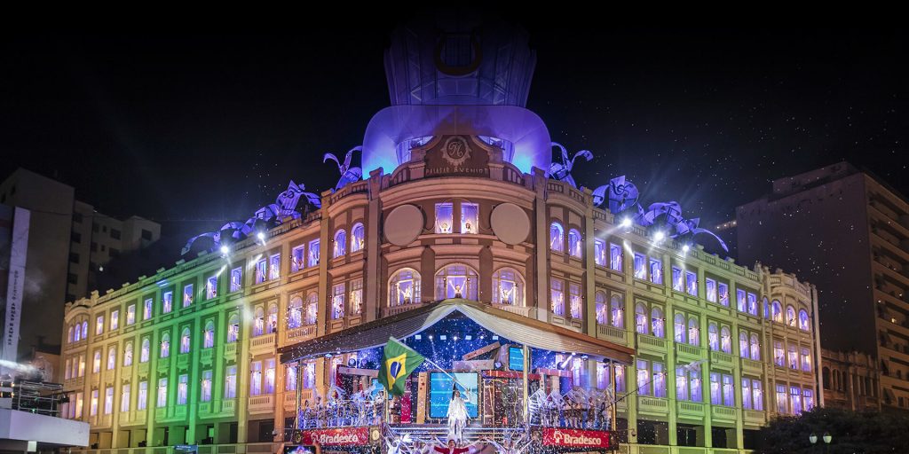 Notícia: Cartão postal de Curitiba completa 90 anos. Foto do palácio avenida de Curitiba em seu tradicional festival de natal. Cores presentes na foto: azul, verde, amarelo, vermelho, bege e marrom.