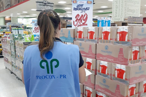 Notícia: Procon-PR fiscaliza preços de produtos em mercados. #PraCegoVer: na foto, uma mulher vestida com um colete azul do Procon do Paraná, realizando inspeção nos mercados. Cores na foto: azul, vermelho, branco, verde, amarelo e laranja. 