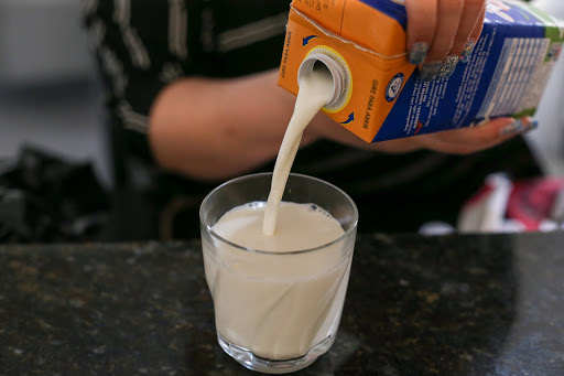 notícia: Procon Notifica Fabricantes de Laticínios Por Aumento no Preço do Leite. #Pracegover: Na foto, uma pessoa colocando o leite em um copo. Cores na imagem: laranja. azul, preto e verde. 