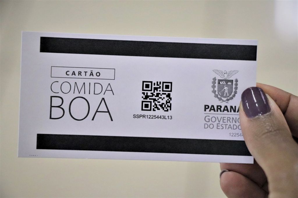 Notícia: Cartão Comida Boa será distribuído nesta terça (26) e quarta (27), em Curitiba. #pratodosverem: na foto,uma mulher segurando o cartão comida boa paraná. Cores na foto: branco, preto e roxo. 