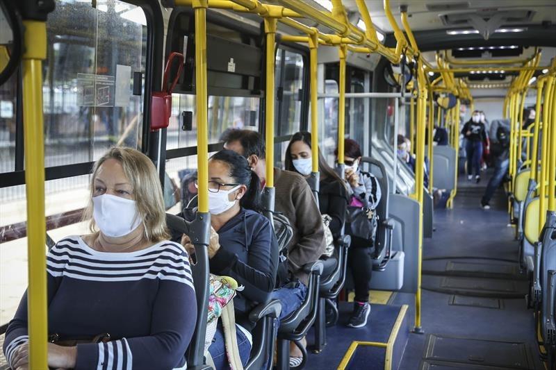 notícia: Horário do comércio é discutido para evitar superlotação em ônibus de Curitiba. #pratodosverem: na foto, um onibus circulando em Curitiba, todos de máscara. Cores na foto: amarelo, preto, cinza, branco, verde.