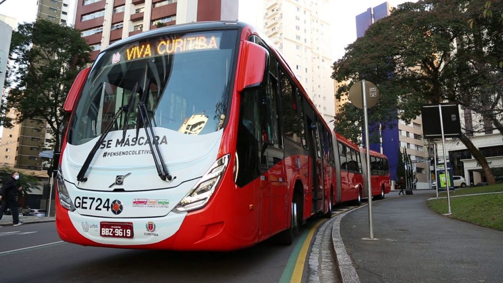 notícia: Só passageiros sentados dura um dia: volta lotação de 50% nos ônibus de Curitiba. #pratodosverem: na foto, o biarticulado de Curitiba. Cores na foto: vermelho, verde, azul, amarelo, preto.